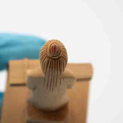 Top-down view of one of the carved wooden figurines of men playing instruments.: Up close top-down image of one of two carved wooden figurines of men playing the Chinese zither. They are carved from light brown cedar wood. There is a small red mark on the bun of the men.