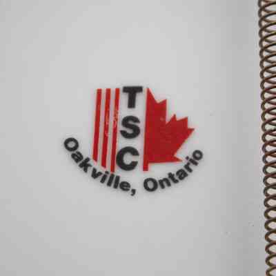 Maker Stamp: Close-up image of the plate's bottom which features the logo of TSC, the plate's maker. It is three red vertical stripes next to TSC in black capital letters, followed by the right half of a red Canadian Maple leaf. Below the logo is "Oakville, Ontario" in black characters.