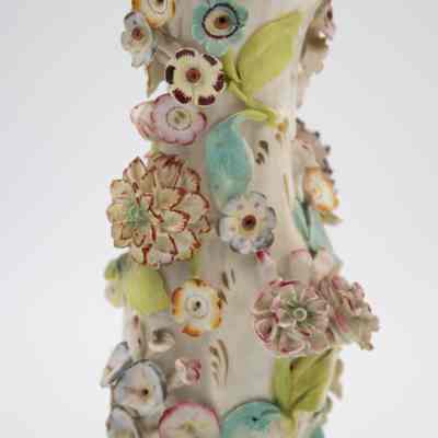 Details on the porcelain bud vases.: Up close image of one of the two porcelain vases decorated with pastel hand-painted encrusted flowers. The flowers are pink, blue, purple, red, and orange with green leaves. There is also gold foil detail around the edges of the vase and throughout the piece.