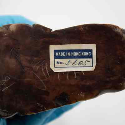 Bottom of the carved stone scene.: Up close image of the bottom of the carved soapstone figurine. It is engraved with Chinese characters and has a white and blue sticker that reads "MADE IN HONG KONG No. 5605."