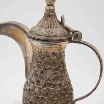 The larger coffee pot from the coffee service given to the Jordans by Sheikh Ahmad Al-Jaber Al-Sabah.: Up close image of the silver debossed coffee pot decorated with vines and flowers and a pointed lid.