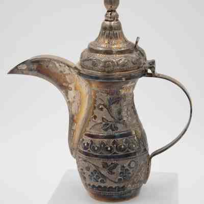 The smaller coffee pot from the coffee service given to the Jordans by Sheikh Ahmad Al-Jaber Al-Sabah.: Up close image of the silver debossed coffee pot decorated with vines and a pointed lid.