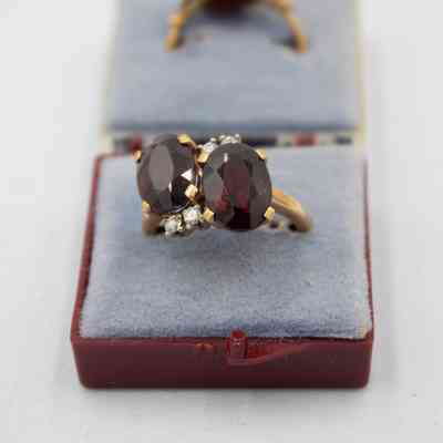 One of the gold, garnet, and diamond rings that belonged to Mrs. Jessie Jordan.: Up close image of one of the gold and garnet rings. It has two large stones with four small diamonds on a thin gold band. It is sitting on a blue cushion in a maroon case.