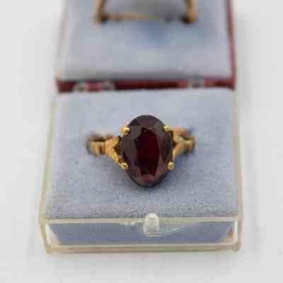 One of the gold, garnet, and diamond rings that belonged to Mrs. Jessie Jordan.: Up close image of one of the gold and garnet rings. It has one round stone in a pronged setting on a thin gold band. It is sitting on a blue cushion in the clear lid of the plastic maroon case.
