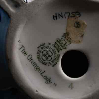 Bottom of the figure.: The bottom of the figure shows the company’s name, logo, and the figures name. “Made in England, Royal Doulton England. “The Orange Lady” H.N. 1759.” The companies logo is a lion with a crown, standing on top of a crown.