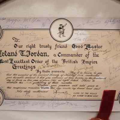 Certificate: Close-up image of the certificate. The image displays a framed certificate addressed to Leland T. Jordan, titled "Good Master and Commander of the Most Excellent Order of the British Empire," in ornate calligraphy. The Society of Falstaff invited Jordan to a meeting on the 14th of June, 1961. The certificate features four circular illustrations at the corners, including a barrel, a plate of food, the head of a boar, and a mug with an overflowing head of beer. The top middle also has a circular illustration and it is of a soldier pointing to the sky. There are several signatures on the certificate, written in the empty beige and white space of the certificate.