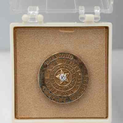 One of L.T. Jordan's Texas A&M Diamond Century Club member pin in white box.: Image of one of L.T.'s Texas A&M Diamond Century Club member pins. The pin is a circle with a gold star and small diamond in the middle on top of a block "T." The pin is approximately an inch in diameter and is debossed with the words "DIAMOND CENTURY MEMBER" in the outer circle and "TEXAS A&M UNIVERSITY" in the inner circle. There is also the olive branches from the Texas A&M seal with the numbers "1876" in the middle. The pin is in a white box with clear lid and sits on a tan felt.