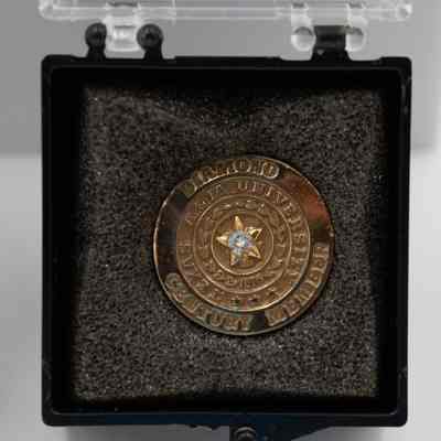 One of L.T. Jordan's Texas A&M Diamond Century Club member pin in black box.: Image of one of L.T.'s Texas A&M Diamond Century Club member pins. The pin is a circle with a gold star and small diamond in the middle on top of a block "T." The pin is approximately an inch in diameter and is debossed with the words "DIAMOND CENTURY MEMBER" in the outer circle and "TEXAS A&M UNIVERSITY" in the inner circle. There is also the olive branches from the Texas A&M seal with the numbers "1876" in the middle. The pin is in a black box with clear lid and sits on black foam.