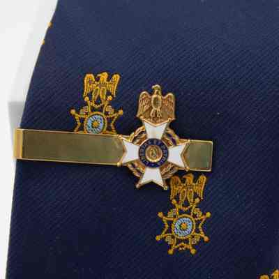 Sons of the American Revolution gold tie clip.: Up close image of the gold Sons of the American Revolution tie clip. The clip has the organization seal attached to the end and has the symbolic eagle, four-point cross, and portrait view of George Washington's head on it. The seal is also enameled with white and blue and reads "LIBERTAS ET PATRIA," the organization's motto.