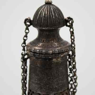 Lid of the silver bottle-shaped decanter with chain-attached lid.: Up close image of the silver bottle-shaped decanter with chain-attached lid. The decanter is engraved with scenes of a Persian Banquet of Song and Dance.