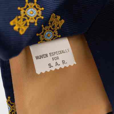 Tag on the inside of the Sons of the American Revolution member tie.: Detailed image of the tag inside the Sons of the American Revolution tie. The tag is white with black print that reads "WOVEN ESPECIALLY FOR S.A.R.". The inside of the tie is a peachy-tan color.
