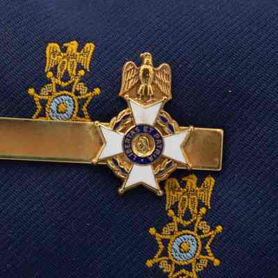 Sons of the American Revolution gold tie clip.: Up close image of the gold Sons of the American Revolution tie clip. The clip has the organization seal attached to the end and has the symbolic eagle, four-point cross, and portrait view of George Washington's head on it. The seal is also enameled with white and blue and reads "LIBERTAS ET PATRIA," the organization's motto.