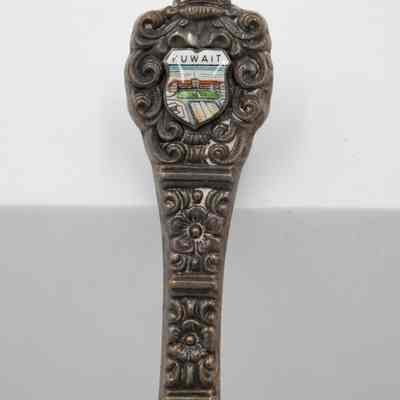 Handle of the letter opener given to the Jordans by His Highness Sheikh Ahmad.: Up close image of the silver handle of the letter opener. It is cast with a floral pattern and the crest at the end has the palace of Kuwait enameled in it. It reads "KUWAIT" in black letters.