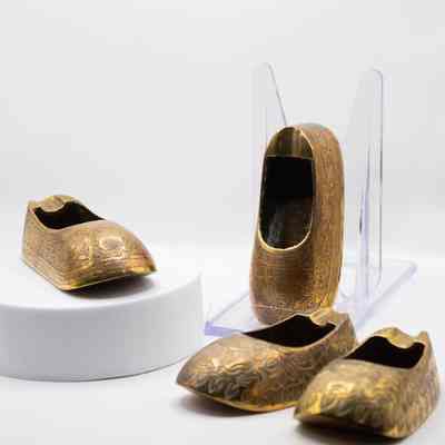 Four shoe ashtrays, the red enameled pair posed with the engraved pair.: The image shows four brass shoe ashtrays featuring intricate red floral designs displayed elegantly on one pair, and an engraved with no enameled pair.
