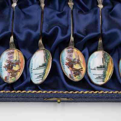 Set of six silver spoons decorated with scenes of Kuwait in a blue case.: Up close image of the alternating designs in the bowls of the spoons. One spoon has a scene of an oil rig system on the bowl. The other design has a camel and Bedouin man in the desert on the bowl. The six spoons are in a blue case.