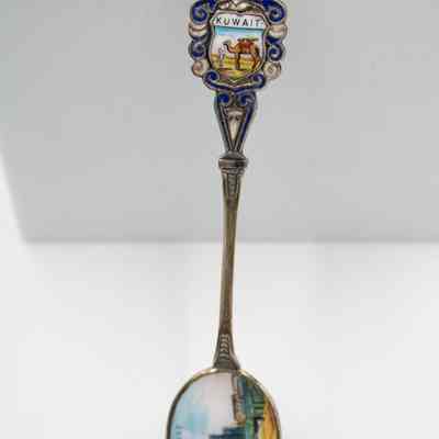 The design of one of the six silver coffee spoons. Three spoons have this design.: Up close image of the enameled design on the handle and bowl of the silver coffee spoon. The scene on the handle depicts a camel and Bedouin man. It is surrounded by blue and white enameled swirls with a knight's helmet at the top. It also reads "KUWAIT." The bowl shows an oil rig scape.