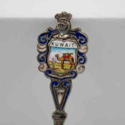 Design on the handle of one of the silver coffee spoons.: Up close image of the enameled handle of one of the silver spoons. The scene depicts a camel and Bedouin man. It is surrounded by blue and white enameled swirls with a knight's helmet at the top and reads "KUWAIT."