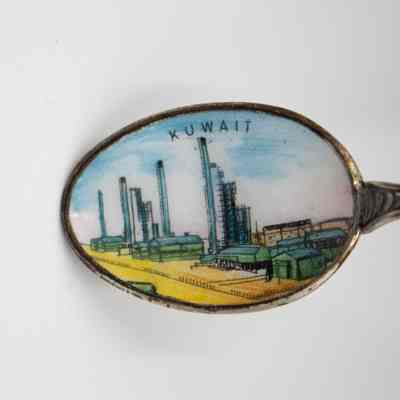 Design in the bowl of a silver coffee spoon.: Up close image of the bowl of one of the silver coffee spoons. The inside of the bowl is enameled with an oil rig scape and reads "KUWAIT" in small black print.