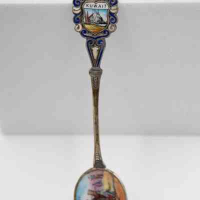 The design of one of the six silver coffee spoons. Three spoons have this design.: Up close image of the enameled design on the handle and bowl of the silver coffee spoon. The scene on the handle depicts an oil rig boat at port. It is surrounded by blue and white enameled swirls with a knight's helmet at the top and reads "KUWAIT." The bowl shows an camel and Bedouin man.