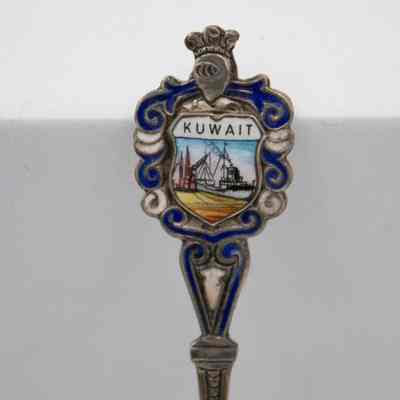 Design on the handle of one of silver coffee spoons.: Up close image of the enameled handle of one of the silver spoons. The scene depicts an oil rig boat at port. It is surrounded by blue and white enameled swirls with a knight's helmet at the top and reads "KUWAIT."