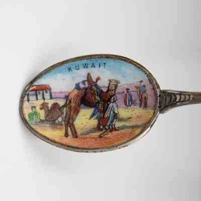 Design in the bowl of a silver coffee spoon.: Up close image of the bowl of one of the silver coffee spoons. The inside of the bowl is enameled with a camel and Bedouin man and reads "KUWAIT" in small black print.