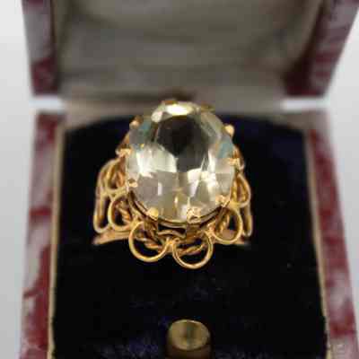 Gold topaz ring that belonged to Mrs. Jessie Jordan.: Up close image of the gold ring with large yellow topaz stone in a 10-pronged setting. The ring is in a red marble plastic box with a blue felt pillow.