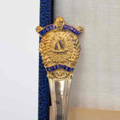 Top of Spoon Handle: Close-up image of the top of the soup spoon handle. It is a gold floral design with a Kuwaiti dhow in the middle. Above it is a blue scroll with the words "LODGE KUWAIT" in gold. There is also a golden crown. Below the dhow is another blue scroll with "N 6810 E.C" in gold. Below this scroll is a small Masonic Crest in gold. The spoon and handle is weathered.