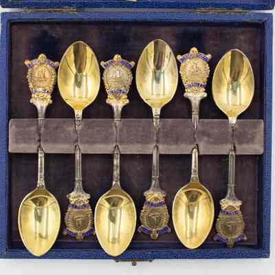 Spoons in Case: Top-down image of the six coffee spoons in the case. The spoons are arranged in opposing directions and are held in place by purple felt. The bowl is gold while the handles are predominantly tarnished silver apart from the golden Masonic Crest ornaments at the top of each handle.