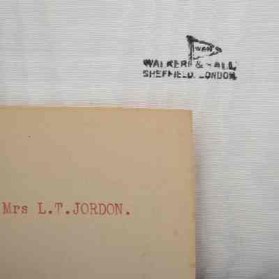 Note and Maker's Signature: Close-up image of the note and the case maker's signature. The note is a yellowed card and in red ink has "Mrs L.T. JORDON. [sic]" printed on it. The case's signature is black and stamped onto the case's white backing is "WALKER & HALL SHEFFIELD LONDON." A small flag with the letters "WAH" is also part of the stamp.