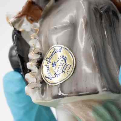 Sticker on the back of the painted porcelain figurine.: Up close image of the gold and blue sticker on the back of the painted porcelain figurine. It reads "Hand Painted WALES CHINA JAPAN."