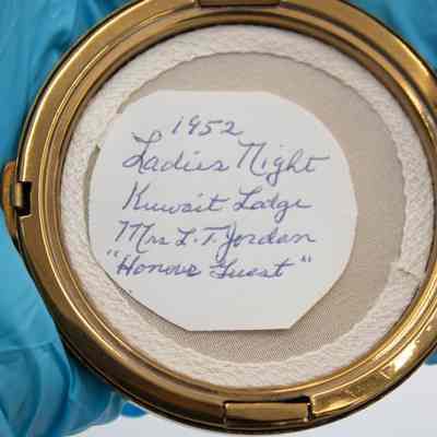 Note found inside the circular gold compact.: Up close image of the note found inside the gold compact. In the face powdered compartment, there is a circular white note that reads "1952 Ladies Night, Kuwait Lodge, Mrs. L.T. Jordan 'Honored Guest'" in blue cursive. Under the note is the remains of the face powder puff. It is white with small woven holes.
