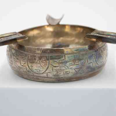 Tiki ashtray: A small silver ashtray features engraved tiki designs and cigarette holders along the rim.