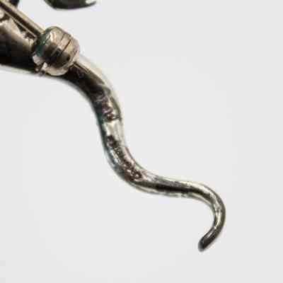 Back of the tail of the small silver lizard brooch.: Up close image of the bottom of the tail of the silver lizard-shaped brooch. It presumed to be engraved with the name of the maker, but it is illegible.
