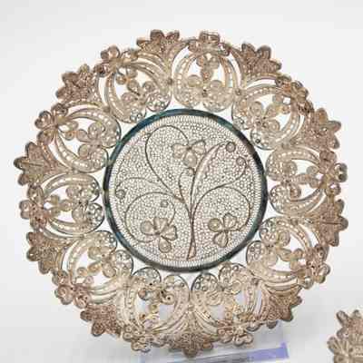 Small round silver filigree lace tray.: Image of one of the round silver filigree lace tray propped up. There is a leaf and floral design around the edge and a three-stemmed plant in the center.