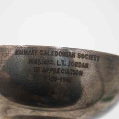 The engraving on the small silver trophy cup presented to the Jordans by the Caledonian Society.: Up close image of the engraving on the outside of the silver trophy cup. The cup is engraved with “KUWAIT CALEDONIAN SOCIETY MR. & MRS. L.T. JORDAN IN APPRECIATION 1949-1961.”