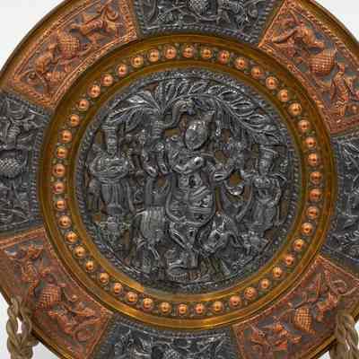 Vishnu plate close-up: A brass orange and black plate. The rim is split into 8 alternating black and orange sections. Each rim section features a relief of two birds eating. The center of the plate is solid black with a relief of Vishnu, a cow and other people.