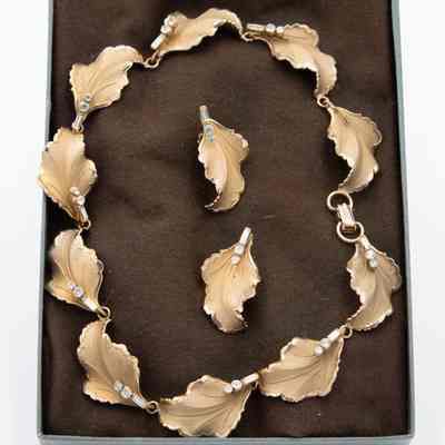 Gold leaf-shaped necklace and earrings with diamonds at each stem, in a box: A gold leaf-shaped necklace and matching earrings adorned with diamonds are displayed in a box on a brown cushion.