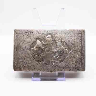 Top of Silver Repousse Box from Persia.: Image of the top of the silver repousse box as it is propped up on a stand. The scene depicts a woman dancing in the center with two people on either side playing instruments. The box is intricately decorated with swirls and leaves. There are small dragon heads on either side of the group.