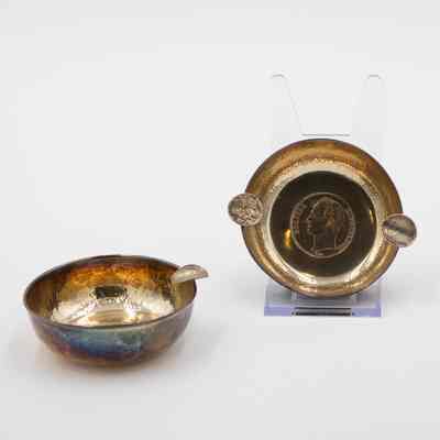 Pair of Small Brass Ashtrays: Image of the two brass ashtrays. The one on the right is on a plastic stand which reveals that the bottom of the ashtray has a Bolivar stamped into its center. The coin takes up much of the bottom and its obverse depicts a bust of Simon Bolivar looking to his left, with the word "Bolivar" on the left and "Libertador" on the right. The two cigarette holders are made out of bent bolivars. The other ashtray is a similar size but has only one cigarette holder.