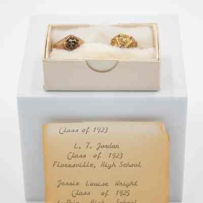 The Jordans' class rings and yellowed note associated with them.: Image of the yellowed note found with the Jordans' class rings. Reads "Class of 1923, L.T. Jordan Class of 1923 Floresville, High school" on the top and then "Jessie Louise Wright Class of 1925 Lufkin High School" on the bottom. It is propped up against a white block. L.T. and Jessie Jordan's class rings sit in white cotton inside a white box. L.T.'s ring is on the left and is bronze with a large "F" in the center with the words "High School" around. Jessie's ring is on the right and is gold. There is a panther head in the center and "L.H.S" around the center.
