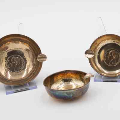 Pair of Small Brass Ashtrays: Image of the two brass ashtrays. The one on the right is on a plastic stand which reveals that the bottom of the ashtray has a Bolivar stamped into its center. The coin takes up much of the bottom and depicts a bust of Simon Bolivar looking to his left, with the word "Bolivar" on the left and "Libertador" on the right. The two cigarette holders are made out of bent bolivars. The other ashtray is a similar size but has only one cigarette holder.