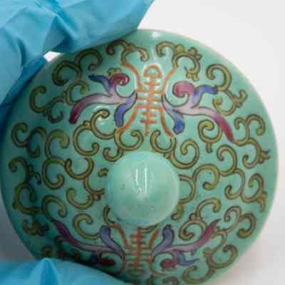 Lid of the mint green porcelain jar.: Up close image of the mint green lid decorated with green swirls and purple, orange, and blue details.