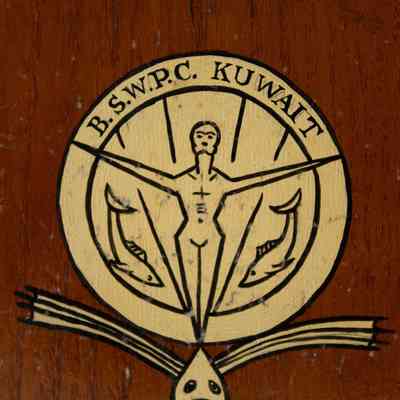 Close up on plaque details: A close up showing the plaques gold circle with a simply drawn man. His arms are up and out in a proud pose. There are two fish near his legs and a forward facing fish with big fins at the bottom. The top of the plaque says "B.S.W.P.C KUWAIT."