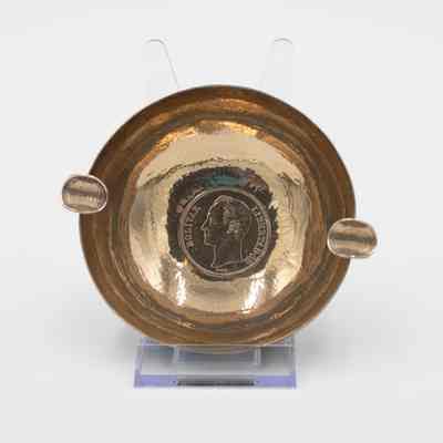 Brass Ashtray: Image of the brass ashtray on a plastic stand to have the interior be visible. The bottom of the ash tray has a Venezuelan Bolivar stamped into its center. The coin depicts the bust of Simón Bolívar facing left with "Bolívar" on the left of the coin and "Libertador" on the right. "Barre" is below Bolívar's bust. There are two cigarette holders on each end of the ashtray, each made with bent Bolivars.