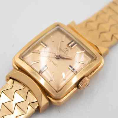 The square watch face of the 18K gold Omega Ladymatic watch given to Mrs. Jessie Jordan by Sheikh Abdullah Mubarak Al Sabah.: Up close image of the gold watch face on the Omega Ladymatic watch. Along with the watch face being gold, so are there watch hands and numerals. There is a small Omega symbol on the small dial on the right side of the face. The glass on the watch face is scratched.
