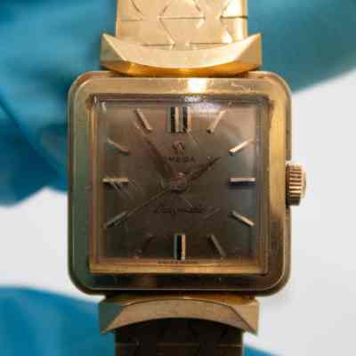 The square watch face of the 18K gold Omega Ladymatic watch given to Mrs. Jessie Jordan by Sheikh Abdullah Mubarak Al Sabah.: Up close image of the gold watch face on the Omega Ladymatic watch. Along with the watch face being gold, so are there watch hands and numerals. The face reads "OMEGA," with the Omega symbol, "Ladymatic" in cursive, and "SWISS MADE" in a small font at the bottom. The glass on the watch face is scratched.