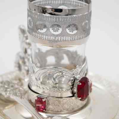 One of the cups, spoons, and saucers from the set.: Up close image of a set of one cup, spoon, and saucer. The cup is glass in a silver plated holder with an etched design and red gemstones. The plate and spoon are also silver plated.