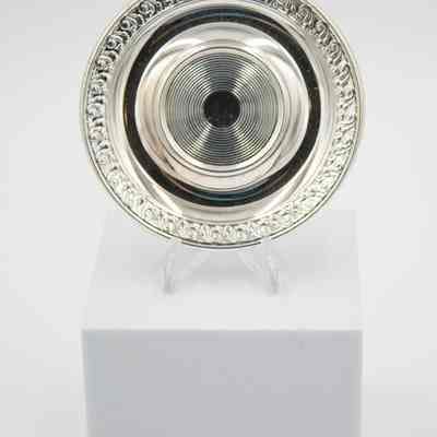 One of the silver plated saucers.: Image of one of the silver plated saucers propped up on a white block. There is a pressed circular design in the center and a leaf and swirl design around the outer edge.