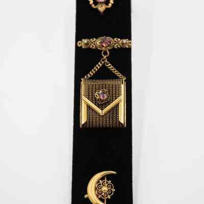 Set of three pins displayed together with Mrs. Jessie Jordan's jewelry collection.: Image of the three pins attached to a black felt display strip. The top pin is a old fleur d'lis with purple amethyst center. The middle pin is a gold pin is set with a garnet-colored stone and two small pearls on either side. Hanging from the bar is a small gold-chain purse with a working clasp; the purse opens and closes but doesn’t hold anything. There is a small gold flower in the center of the purse with an purple amethyst center. The bottom pin is a crescent moon shape with a gold flower in the center.