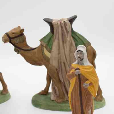 One set of the camel and male Bedouin figurine.: Image of one of the male figurines wearing a brown traditional Kuwaiti dresses, a yellow shall, and white head covering with a walking stick. The camel has a yellow and green saddle pad, a large brown saddle, and draped beige cloth. The reins are brown. The male figurine is standing in front of a the camel.